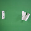 Two Optimum 4000 white metal wall brackets are mounted on a green wall; the right bracket is open at an angle, while the left one is closed and flat against the wall.