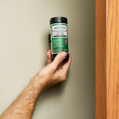 A hand holds a container of Heavy-Duty Stand Alone LB (Leave Behind) air treatment, placing it against a beige wall beside a wooden edge.