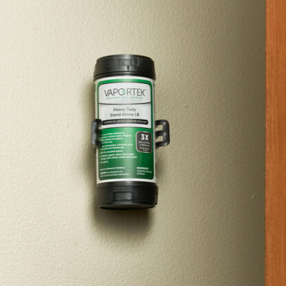 A Heavy-Duty Stand Alone LB (Leave Behind) air freshener canister is mounted vertically on a beige wall near a wooden doorframe, displaying its green and white label.