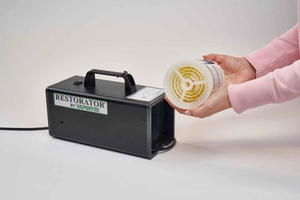 A person holds a cylindrical cartridge near the open compartment of a black machine labeled Restorator by Vaportek, which is sitting on a white surface.