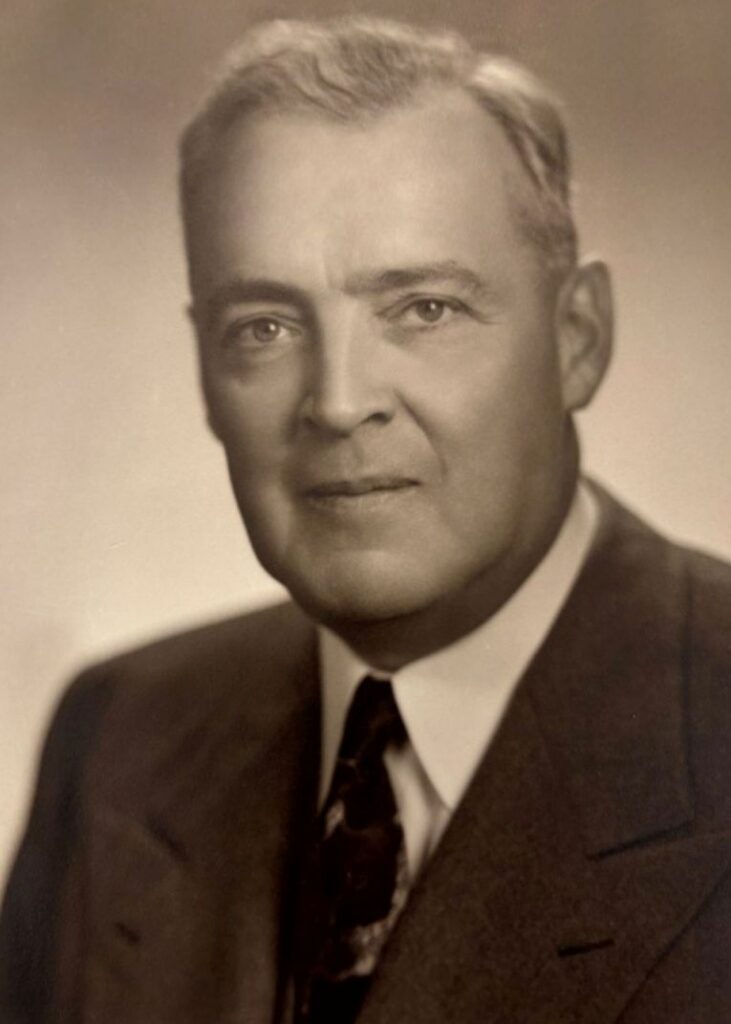 A sepia-toned portrait of an older man in a suit and tie, looking at the camera with a neutral expression. The background is plain and softly blurred.