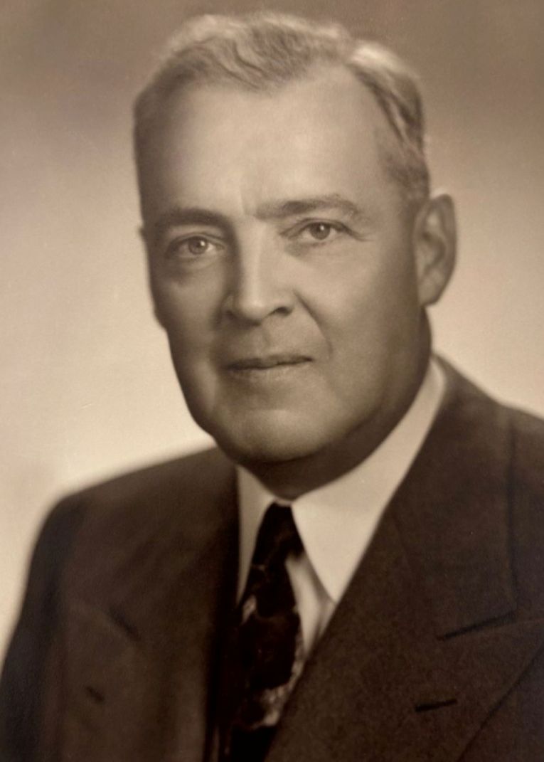 A sepia-toned portrait of an older man in a suit and tie, looking at the camera with a neutral expression. The background is plain and softly blurred.